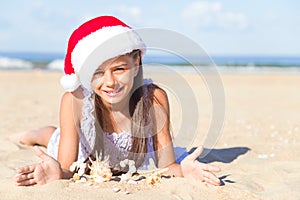 Happy cute smiling little girl in a red Santa hat lying on a sand on a sea beach and showing numbers 2020 made from shells