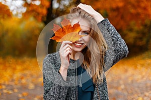 Happy cute young attractive woman with a smile in a fashionable coat is standing in the park and covering her face