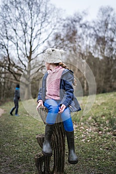 Happy cute young girl in wellington boots sat in autumn countryside looking back away from camera at boy exploring nature