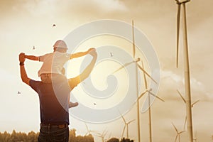 Happy dad carrying son on shoulders checking future project at wind farm site on sunset. Silhouette of father and son