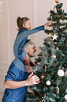 Happy dad and daughter decorate the Christmas tree. A little girl is sitting on Daddy& x27;s shoulders, holding a toy on the