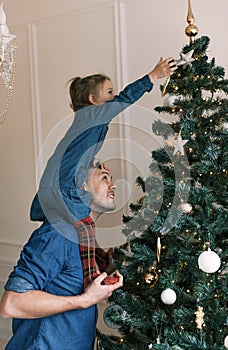 Happy dad and daughter decorate the Christmas tree. A little girl is sitting on Daddy& x27;s shoulders, holding a toy on the