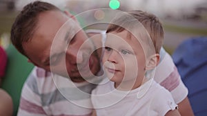 Happy dad kisses on the cheek his cute son. Father and son spending time at the park in summer, boy points to something.