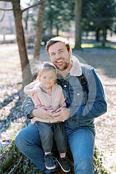 Happy dad with a little girl on his knees sit on a stump in a spring park