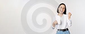 Happy dancing woman drinking coffee or tea from mug. Korean girl with cup, standing over white background