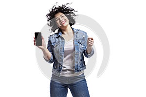 Happy dancing young beautiful girl in jeans shows a smartphone that is emitted on a black screen. Bright brunette with curly hair