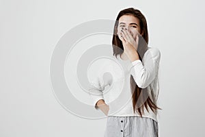 Happy dark-haired girl closing mouth with hand going to see surprise prepared by boyfriend standing and smiling in