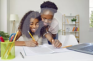 Happy dark-skinned mother watching her cheerful teenage daughter write and draw at home.