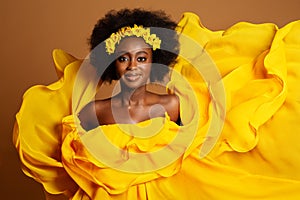 Happy Dark Skinned Woman in Yellow Fashion Dress. Beauty Afro American Model dancing in Silk Gown waving Flying on Wind over Beige