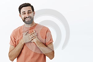 Happy delighted and grateful adult european man with beard in pink t-shirt holding palms on heart and smiling thankful