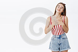 Happy delighted and satisfied attractive female in striped top holding hands on heart from heartwarming feeling in soul