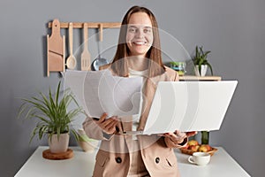 Happy delighted woman with brown hair wearing beige jacket standing in home kitchen interior using laptop computer holding papers
