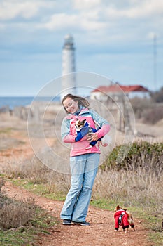 Happy delightful active young woman holding little chihuahua dog in animal clothes near ocean coast with lighthouse