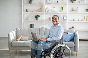 Happy disabled black man in wheelchair using laptop, working online from home, copy space