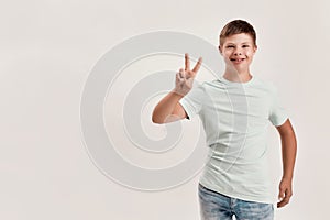 Happy disabled boy with Down syndrome smiling at camera, showing peace sign with one hand while standing isolated over