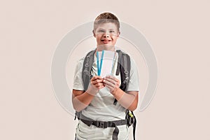 Happy disabled boy with Down syndrome wearing backpack smiling while holding air tickets, standing isolated over white