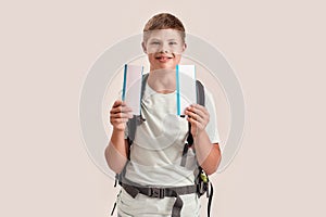 Happy disabled boy with Down syndrome wearing backpack smiling while holding two air tickets, standing isolated over