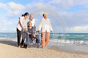 Happy disabled senior woman in wheelchair spending time with her friends on tropical sand beach. Group of four cheerful Asian