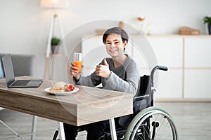 Happy disabled teenager having breakfast, holding glass of juice, showing thumb up gesture at home