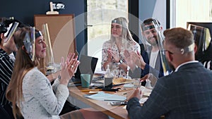 Happy diverse business people team applauding male boss at office meeting, all wearing COVID-19 safety face shields.