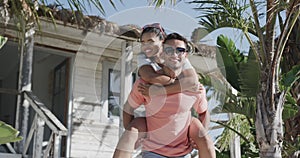 Happy diverse couple having fun piggybacking outside wooden beach house in the sun, in slow motion