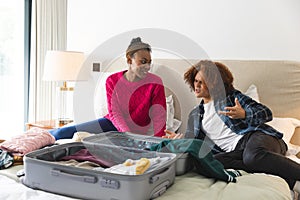 Happy diverse couple lying on bed, talking and packing suitcase in sunny bedroom, at home