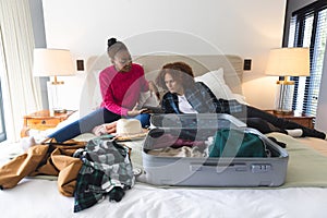 Happy diverse couple lying on bed, using tablet and packing suitcase in sunny bedroom, at home