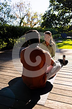 Happy diverse couple practicing yoga sitting on deck smiling in sunny garden, copy space