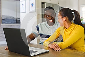 Happy diverse couple using laptop smiling at each other in kitchen