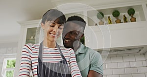 Happy diverse couple wearing blue apron embracing and cooking in kitchen