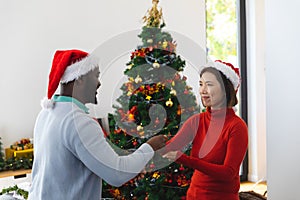 Happy diverse couple wearing santa claus hats, dancing next to christmas tree