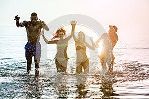 Happy diverse culture friends having fun inside water on the beach - Young people dancing and jumping in sea with back sun light