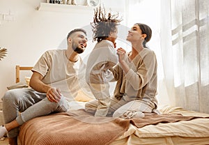 Happy diverse family mom, dad and child  laughing, playing and jumping   in bed   at home