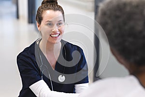 Happy diverse female doctor talking with senior female patient in wheelchair in hospital corridor