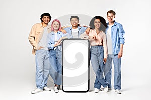 Happy diverse friends posing near big cellphone with white blank screen, showing place for ad, mockup, banner