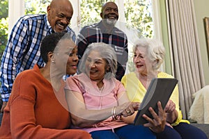 Happy diverse group of senior friends using tablet and laughing in sunny living room