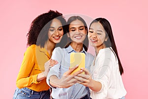 Happy diverse ladies making selfie on cellphone, standing together over pink studio background