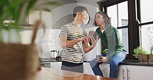 Happy diverse male couple drinking coffee and using smartphone in kitchen