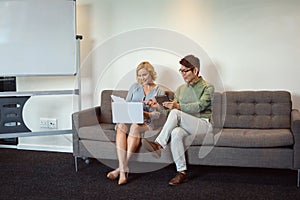 Happy diverse male and female colleague talking in office lounge using tablet and laptop