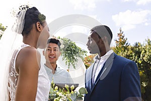 Happy diverse male officiant with bride and groom at outdoor wedding ceremony in sunny garden
