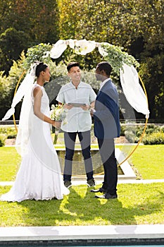 Happy diverse male officiant, bride and groom at outdoor wedding ceremony in sunny garden