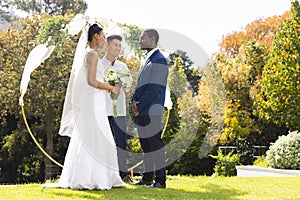 Happy diverse male officiant, bride and groom at outdoor wedding in sunny garden, copy space