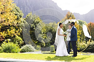 Happy diverse male officiant, bride and groom at outdoor wedding in sunny garden, copy space