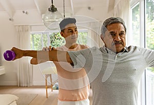 Happy diverse male physiotherapist advising and determined senior male patient using dumbbells