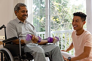 Happy diverse male physiotherapist advising and senior male patient in wheelchair using dumbbells