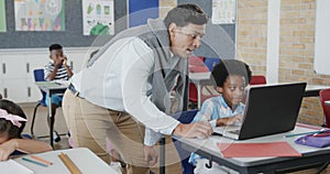 Happy diverse male teacher helping boy at desk using laptop in elementary school class, slow motion