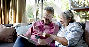 Happy diverse senior couple sitting on couch and using tablet for online shopping at home