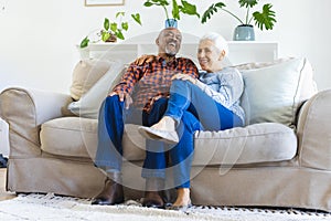 Happy diverse senior couple sitting on sofa, talking and embracing in sunny living room