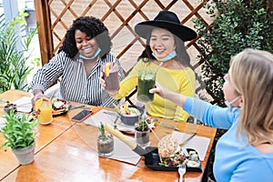 Happy diverse young friends eating at brunch restaurant while wearing safety masks - Focus on asian girl face