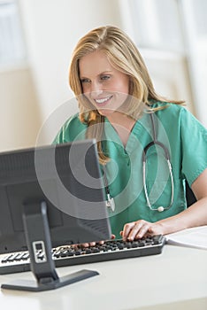 Happy Doctor In Scrubs Using Computer At Hospital Desk